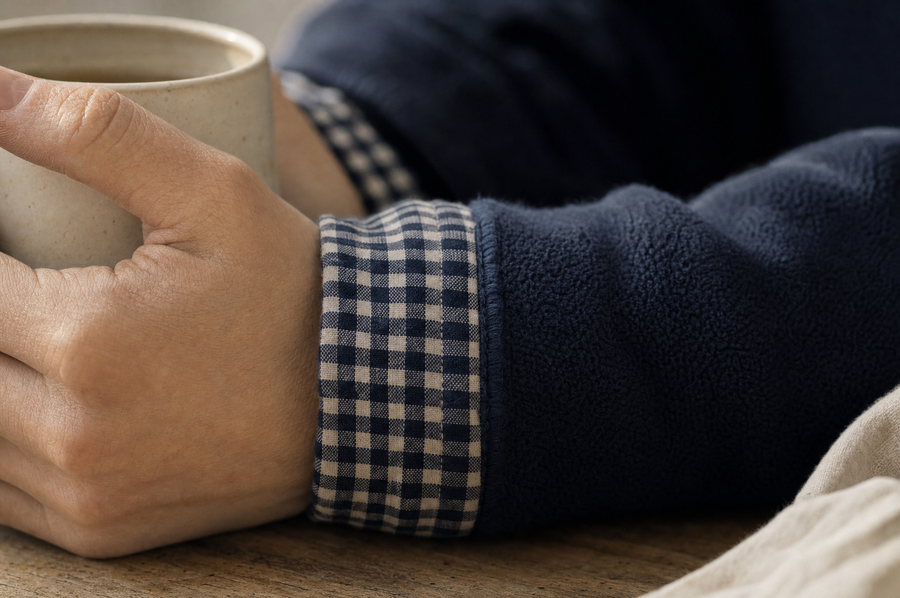 Close-up of a gingham cuff turned back over a navy fleece sleeve, hands around a stoneware mug