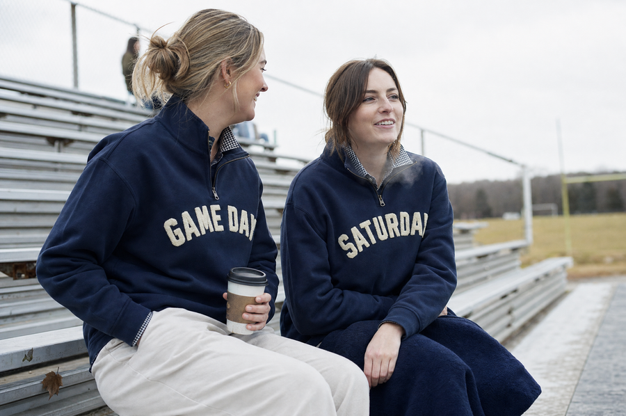 Two friends on bleachers in GAME DAY and SATURDAY quarter-zips on a cold bright afternoon