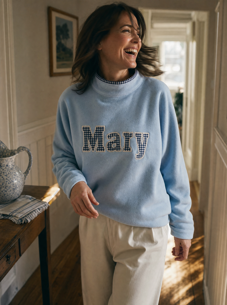 A woman laughing mid-movement in a pale blue sweatshirt with Mary in gingham applique letters