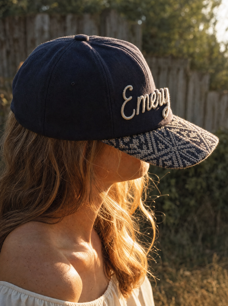 Side profile of a woman in an embroidered cap reading Emery, with a woven tapestry brim, in low sun
