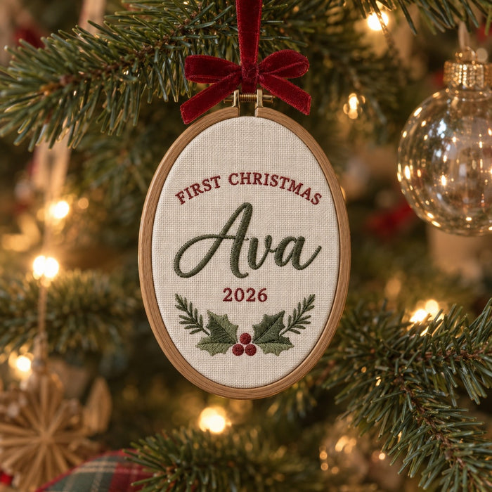 Machine-embroidered First Christmas ornament hoop with the name Ava, the year 2026 and a holly sprig, hanging on a Christmas tree